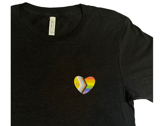 Zoomed in on a black t-shirt is laid flat against a white background. A heart shape rainbow flag is placed over the left chest.